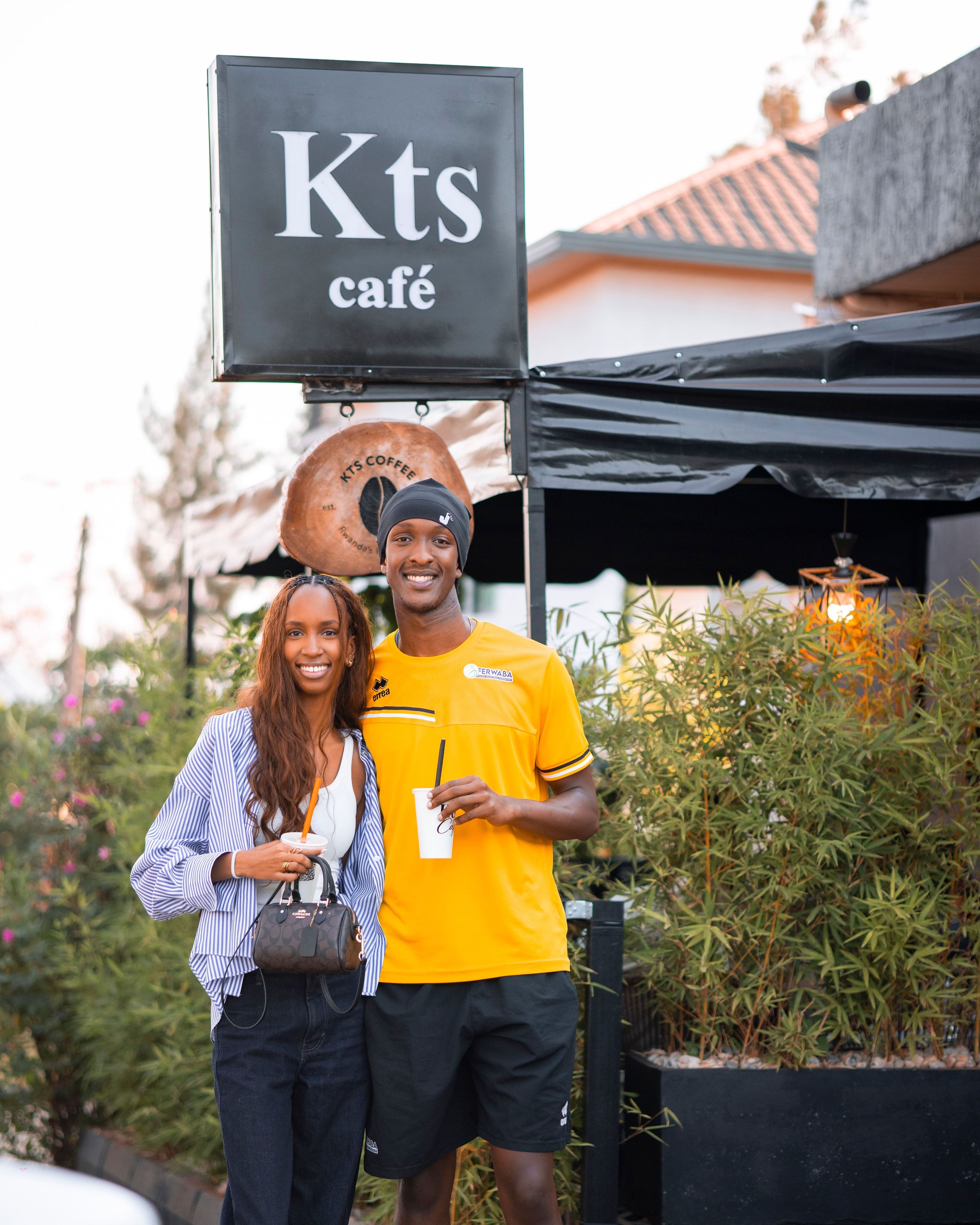 Two smiling visitors holding KTS Café cups under the KTS Café street sign in Kibagabaga