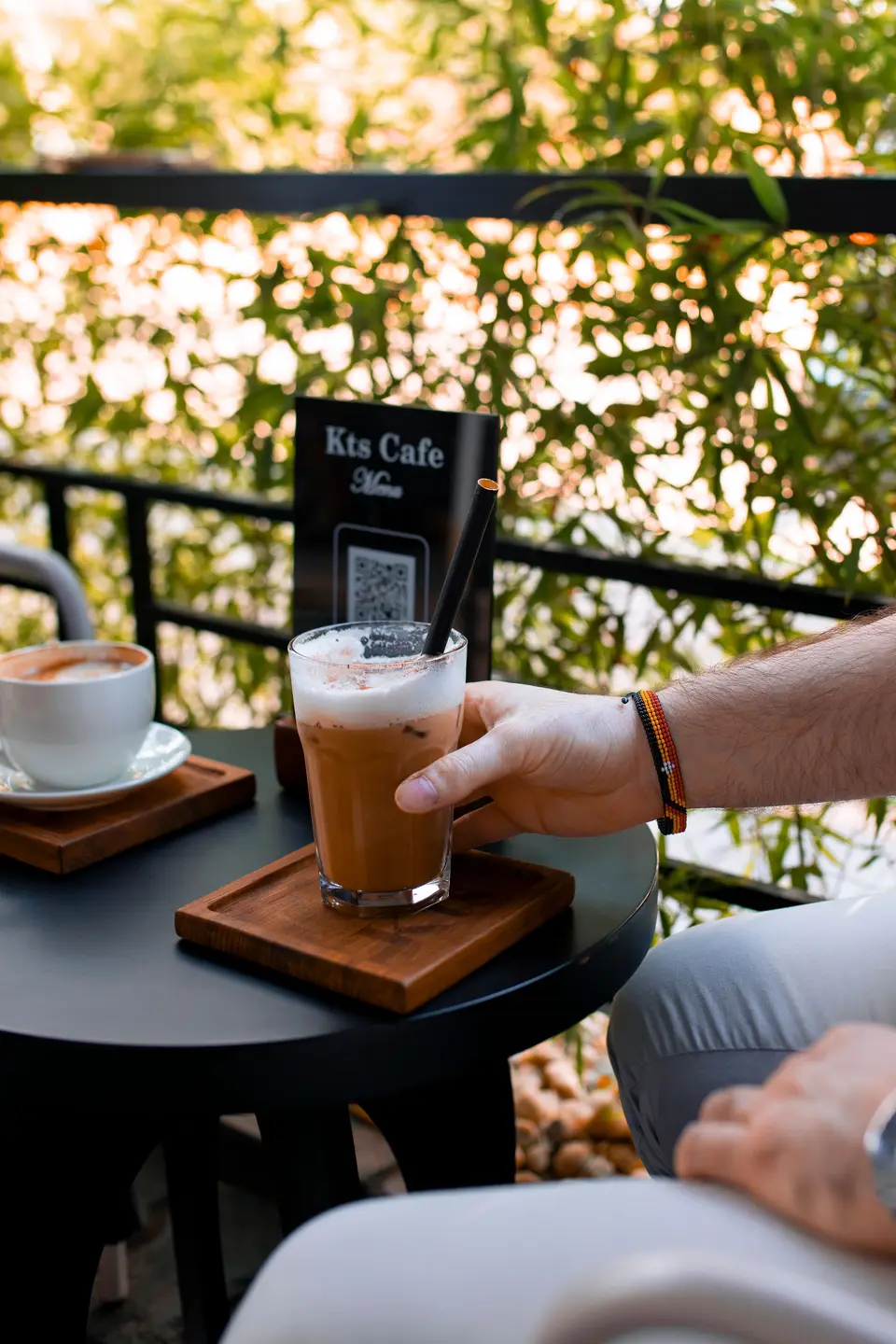 A hand lifting a foam-topped iced latte from its wooden coaster on the KTS Café terrace